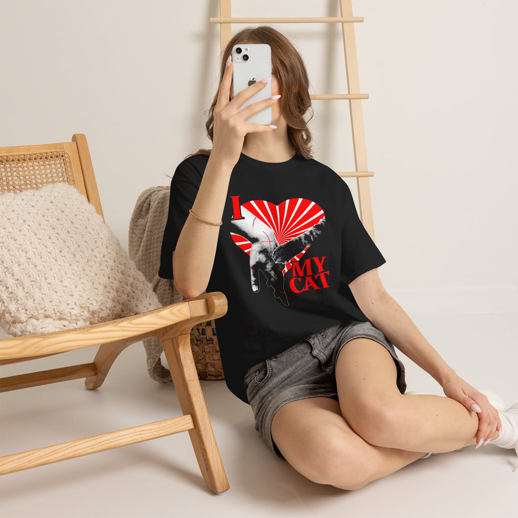 A graphic tee on a sitting lady displaying a funny classical art parody of a scratch-marked human hand and a cat paw over a red heart, reading 'I MY LOVE CAT' in a bold vintage font.